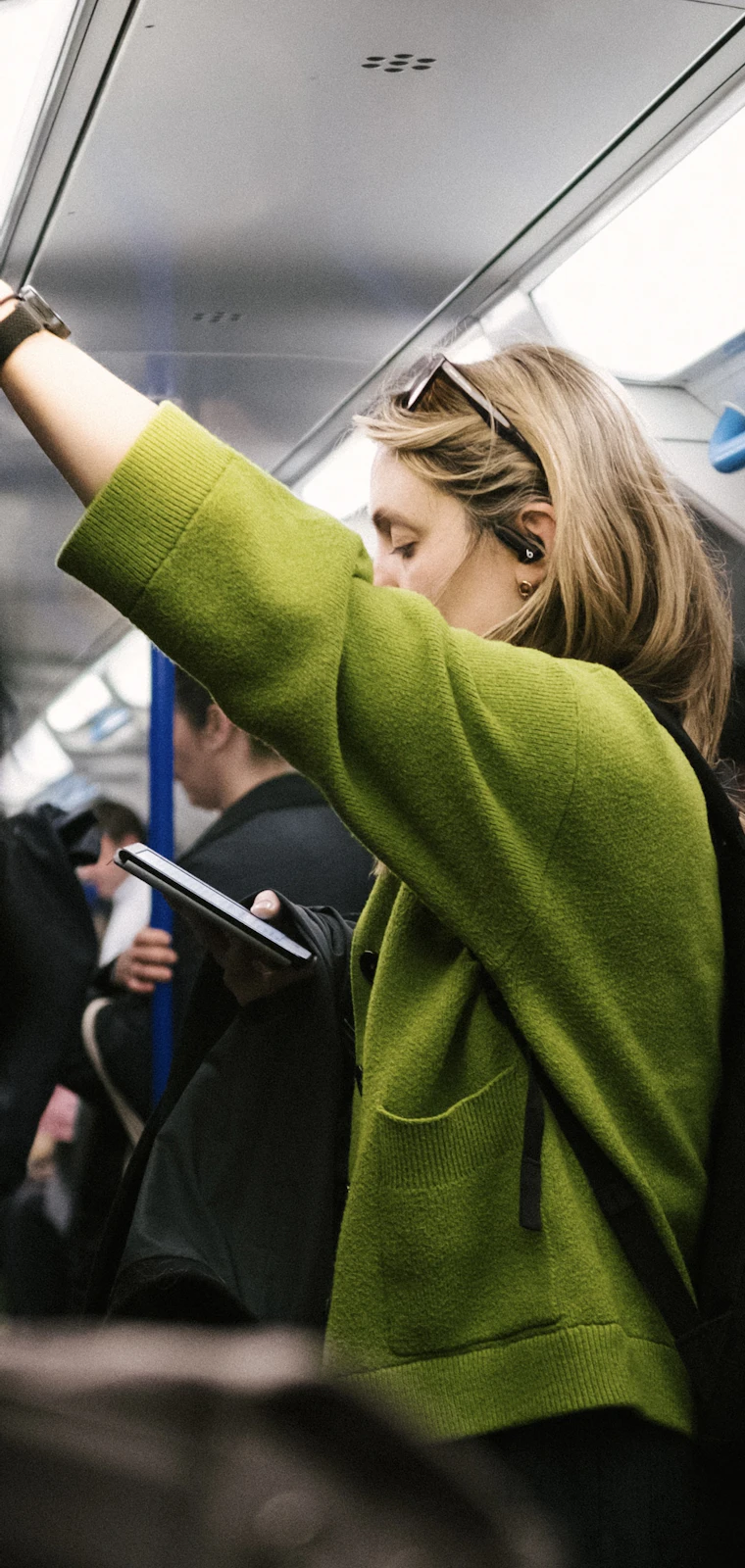 Woman in a green cardigan holding a rail on a crowded train while looking at her smartphone