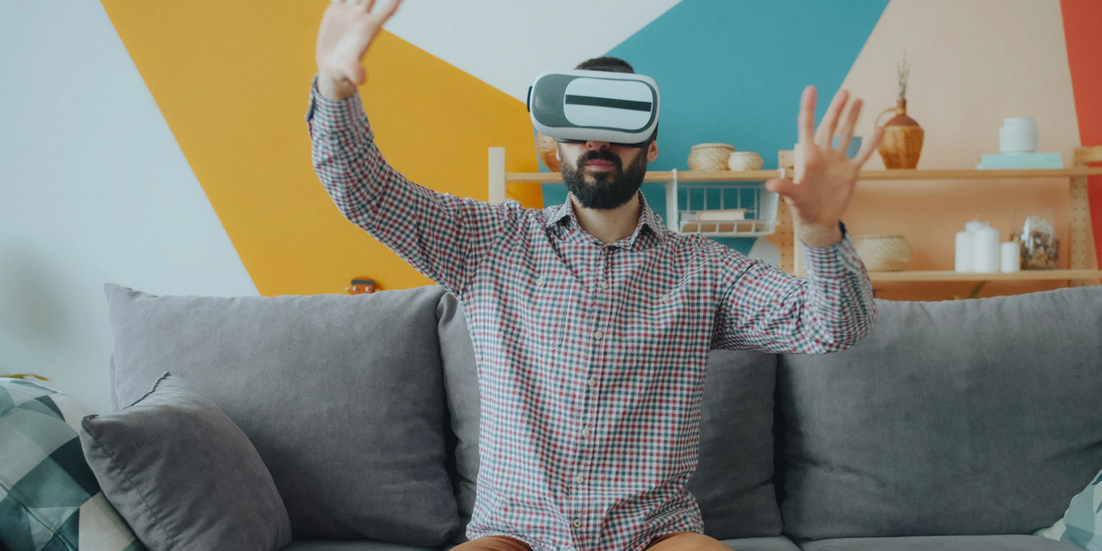 Bearded man in a checked shirt wearing a VR headset on a grey sofa, reaching out with both hands in a colourful living room